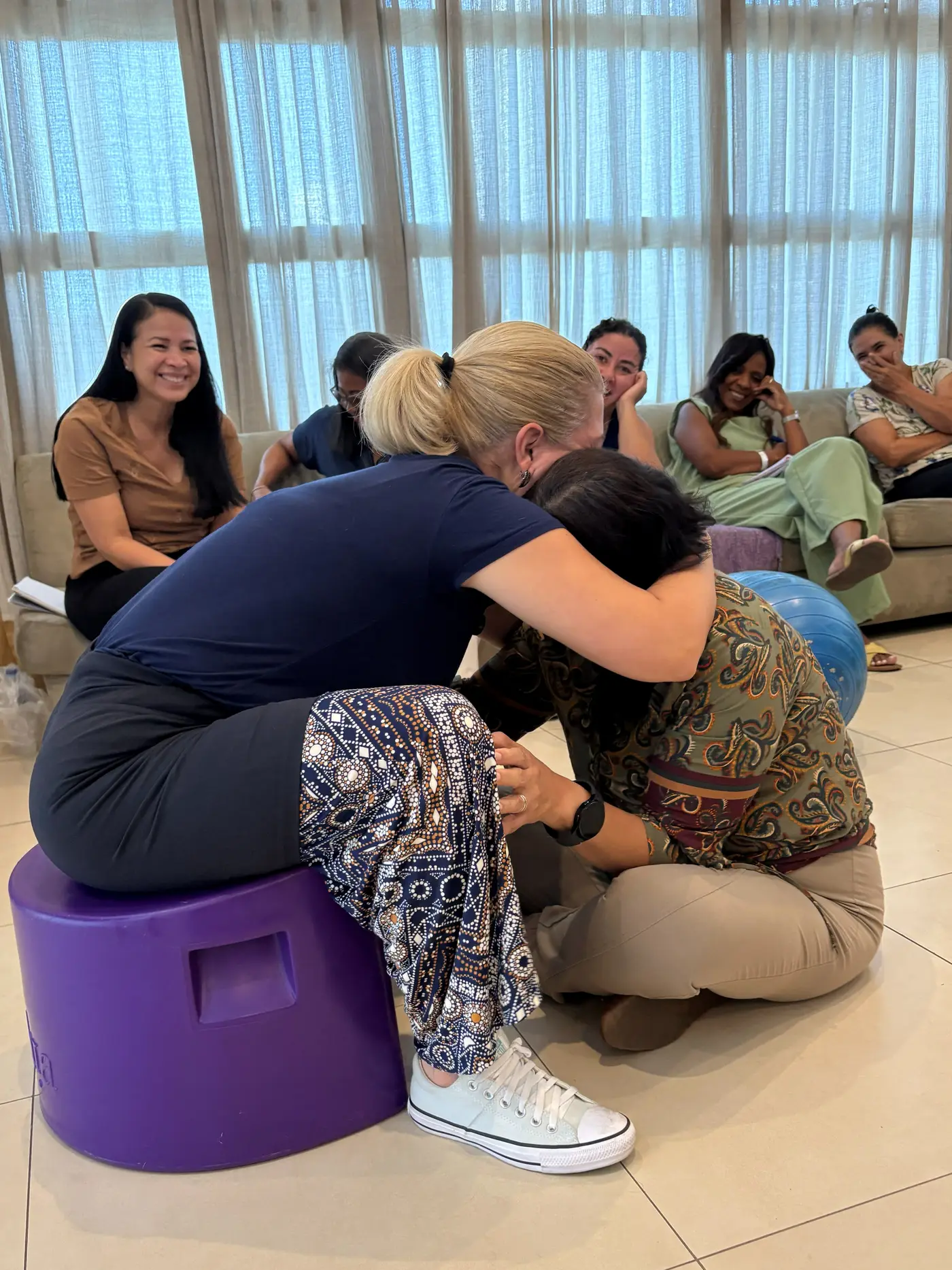 Participante sentada na banqueta de parto recebe apoio de outra por trás, enquanto uma terceira se agacha à frente e a turma acompanha em volta.