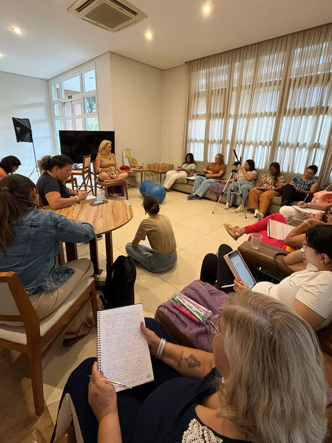 A turma em roda, sentada em sofás e no chão, com a docente à frente e uma bola de parto no centro da sala.