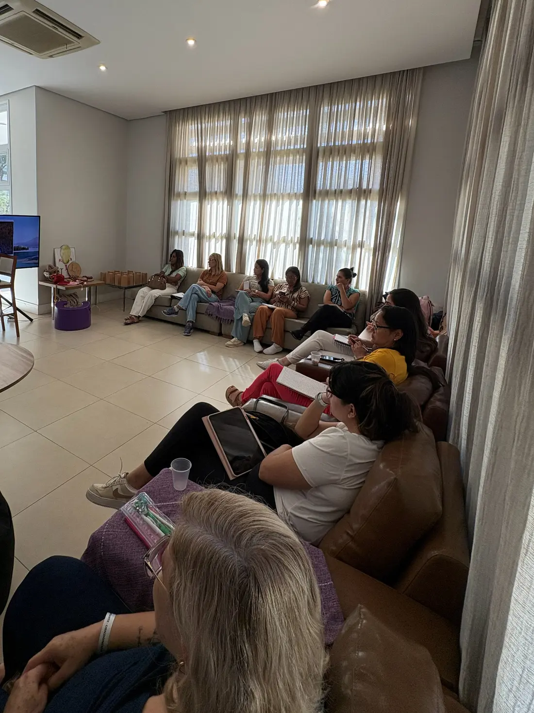 Participantes sentadas em volta da sala, com cadernos e tablets no colo, acompanhando a apresentação na tela.